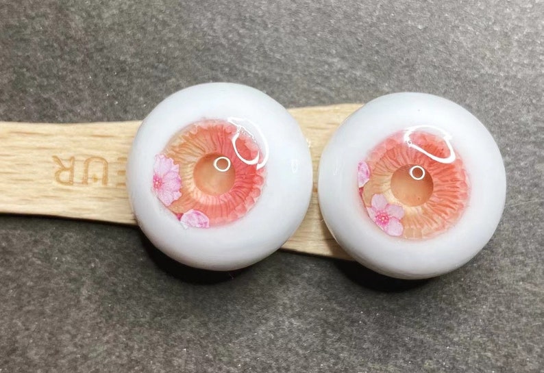 Pode incluir: Dois olhos de boneca brancos com &iacute;ris rosa e p&ecirc;ssego e um design floral. Os olhos est&atilde;o sobre uma superf&iacute;cie de madeira.