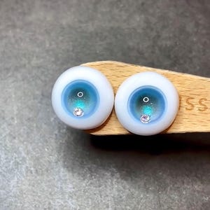 Pode incluir: Dois olhos de boneca azuis e brancos com um pequeno strass transparente no centro de cada &iacute;ris.