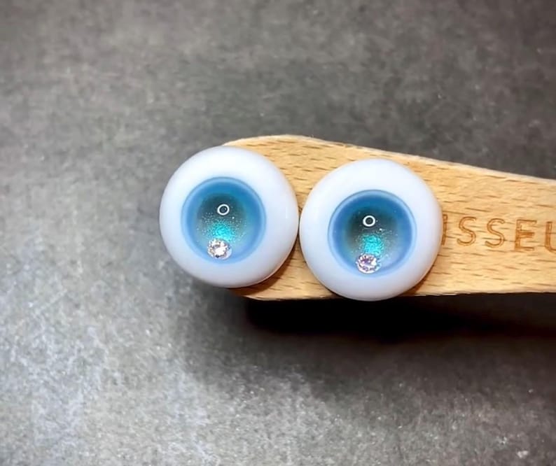 Pode incluir: Dois olhos de boneca azuis e brancos com um pequeno strass transparente no centro de cada &iacute;ris.