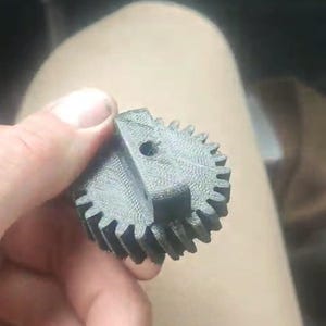 Könnte beinhalten: Ein dunkelgraues, 3D-gedrucktes Zahnrad mit einer kreisförmigen Basis und einer Reihe gleichmäßig verteilter Zähne am Umfang. Auf der Oberseite befindet sich ein erhöhter rechteckiger Abschnitt mit einem kleinen, mittigen Loch.