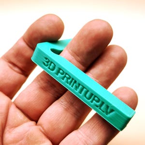 Könnte beinhalten: Ein tealblaues, 3D-gedrucktes Objekt in Form eines Hakens mit dem aufgedruckten Text "3D PRINTUP.LV".