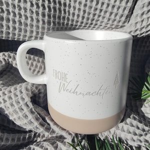 Könnte beinhalten: Ein weißer Keramikbecher mit gesprenkelter Oberfläche und einem hellbraunen Boden. Der Becher trägt den Schriftzug "FROHE Weihnachten" in grauer Schreibschrift, ergänzt durch ein kleines, schlichtes Umriss-Symbol eines Tannenbaums rechts.