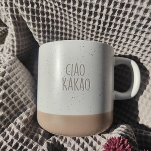 Könnte beinhalten: Ein Keramikbecher mit einem gesprenkelten weißen oberen Teil und einem einfarbigen beigen Boden. Der Text "CIAO KAKAO" ist in der Mitte des weißen Bereichs eingeprägt. Der Becher hat einen geschwungenen Henkel und eine glatte, matte Oberfläche.