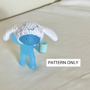 Puede incluir: Un gorro blanco de crochet con orejas de conejo, usado por una figurita azul que sostiene un pequeño objeto azul claro. El texto "PATTERN ONLY" es visible debajo de la figurita.