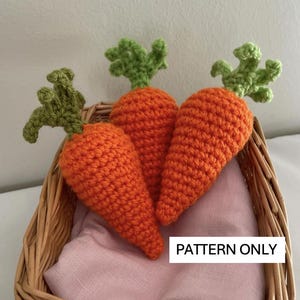May include: Three crocheted carrots in a wicker basket. The carrots are orange with green tops. The text "PATTERN ONLY" is visible in the image.