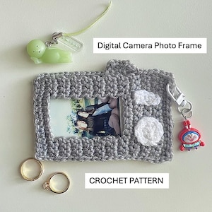 May include: A gray crocheted digital camera photo frame with a photo inside. The frame has white crocheted details and a small red charm. The text "Digital Camera Photo Frame" and "CROCHET PATTERN" are visible.