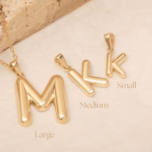 Może przedstawiać: Złoty naszyjnik z wisiorkiem z inicjałem, z dużym wisiorkiem "M" i dwoma mniejszymi wisiorkami "K". Wisiorki są oznaczone jako "Large", "Medium" i "Small". Łańcuszek naszyjnika jest również złoty.