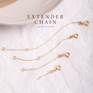 May include: Four gold extender chains of varying lengths, each with a lobster clasp and jump rings. The chains are on a white textured surface. The text "EXTENDER CHAIN 14K SOLID GOLD" is printed above the chains.