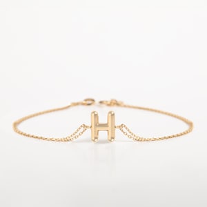 May include: A gold bracelet with the letter "H" as a central charm. The bracelet has a fine chain design and a lobster clasp closure. The letter is in a bold, sans-serif font. The jewellery is set against a white background.
