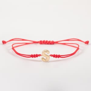 Könnte beinhalten: Ein rotes Armband mit einem goldfarbenen Buchstaben "S" als Anhänger. Das Armband ist mit einem verstellbaren Schiebeknoten versehen. Der Buchstabe ist etwa 1,3 cm hoch. Das Armband wird vor weißem Hintergrund gezeigt.