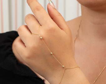 May include: A delicate gold hand chain bracelet. The bracelet features a chain that connects to a ring and a chain that wraps around the wrist. Small gold beads are evenly spaced along the chains. The jewelry is worn on a hand.