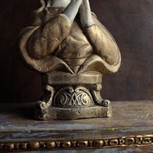 May include: A gold and white painted bust of a person with their hands clasped in prayer. The bust is on a decorative base with intricate details.