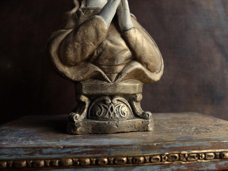 May include: A gold and white painted bust of a person with their hands clasped in prayer. The bust is on a decorative base with intricate details.