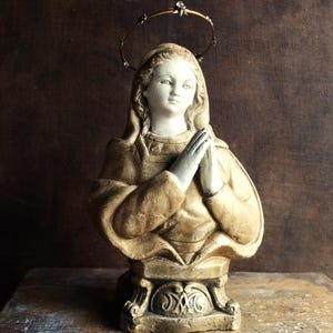 May include: A gold-painted plaster bust of a woman with a halo, wearing a robe and praying with her hands clasped together. The bust is on a pedestal with ornate details.