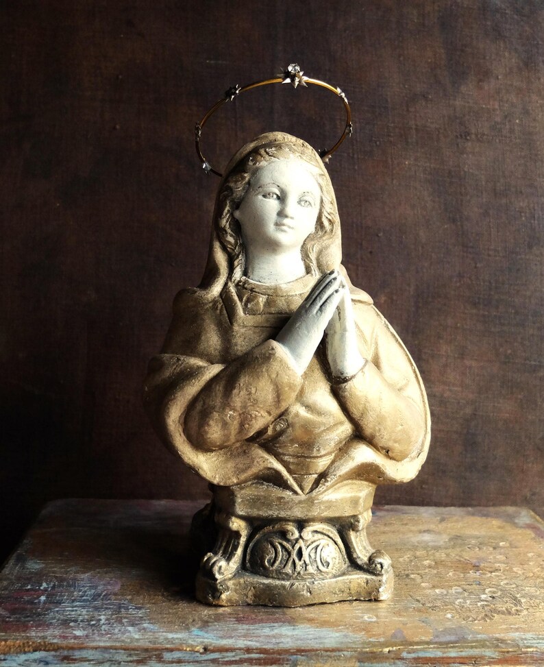 May include: A gold-painted plaster bust of a woman with a halo, wearing a robe and praying with her hands clasped together. The bust is on a pedestal with ornate details.