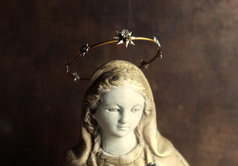 May include: A white plaster statue of a woman wearing a gold halo with star-shaped accents. The statue is wearing a white robe and has a soft, gentle expression.