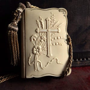 Puede incluir: Estuche en forma de libro de color marfil con un diseño en relieve de cruz y flores. El estuche tiene una cadena y borla doradas. La cubierta presenta una cruz, trigo y flores. El estuche se cierra con un pequeño cierre.