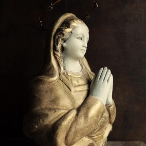 May include: A white plaster statue of a woman with a gold-painted robe and a crown of thorns. She is praying with her hands clasped together.