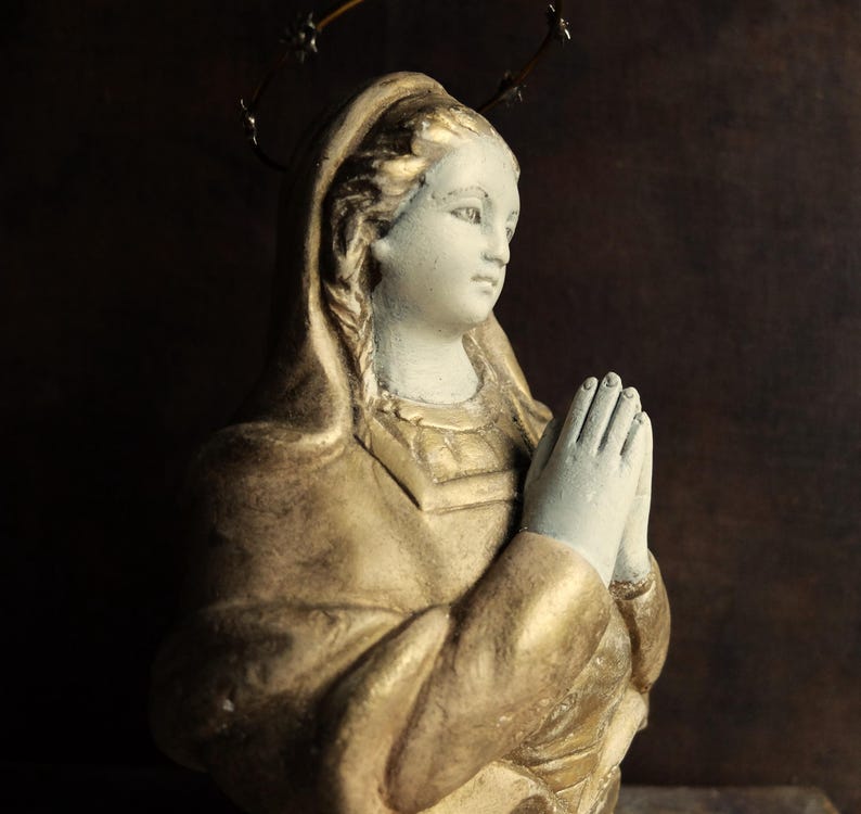 May include: A white plaster statue of a woman with a gold-painted robe and a crown of thorns. She is praying with her hands clasped together.