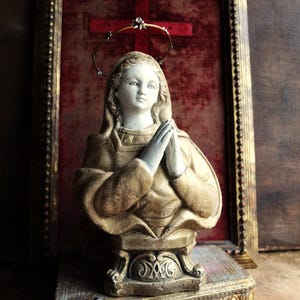 May include: A gold-painted plaster bust of a woman with a crown of thorns halo, praying with her hands clasped together. The bust is displayed on a wooden base with gold accents.