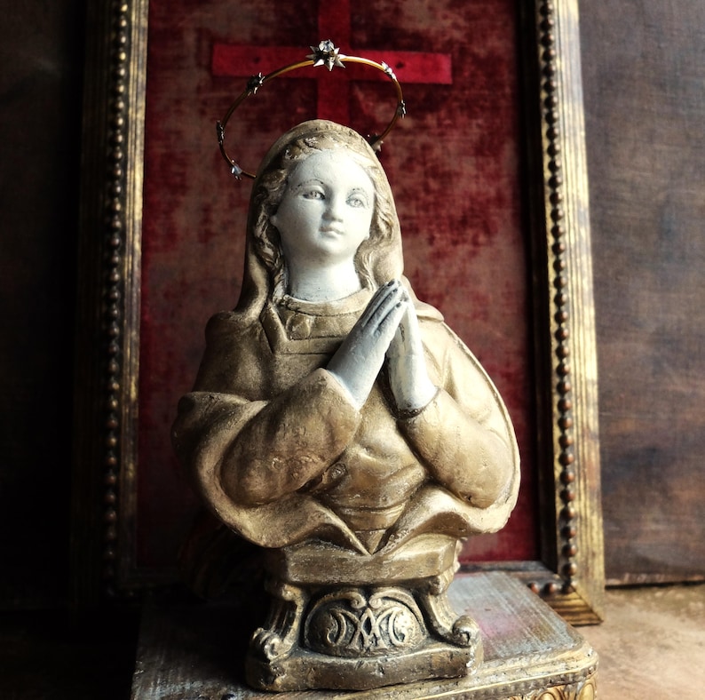 May include: A gold-painted plaster bust of a woman with a crown of thorns halo, praying with her hands clasped together. The bust is displayed on a wooden base with gold accents.