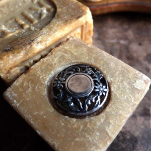 Puede incluir: Primer plano de dos jabones cuadrados de color marrón claro. Un jabón tiene un adorno decorativo de metal plateado con un diseño floral. El otro jabón tiene un diseño en relieve. Los jabones están sobre una superficie oscura.