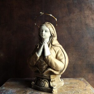 May include: A gold-painted plaster statue of a woman with a halo, wearing a robe and praying with her hands clasped together.