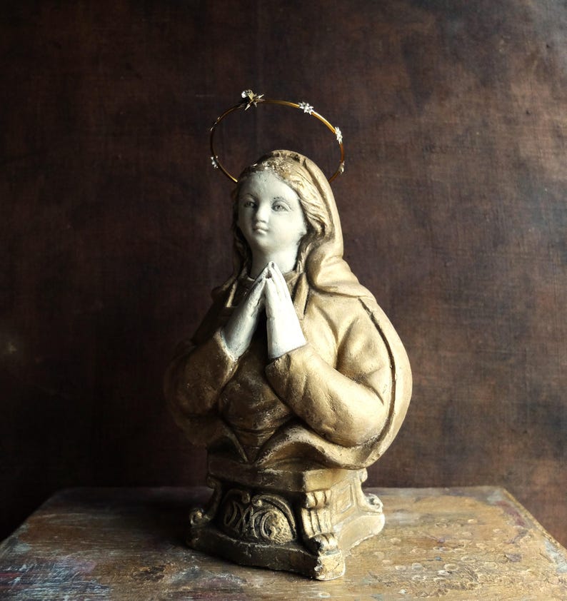 May include: A gold-painted plaster statue of a woman with a halo, wearing a robe and praying with her hands clasped together.