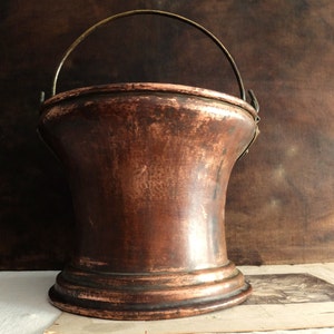 Può includere: Un secchio di rame vintage con un manico in ottone. Il secchio ha una finitura martellata e una base larga e svasata.