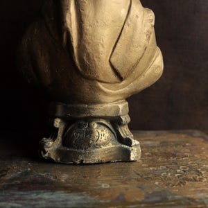 May include: A gold-colored statue of a person with a draped cloth, standing on a pedestal with a decorative base.