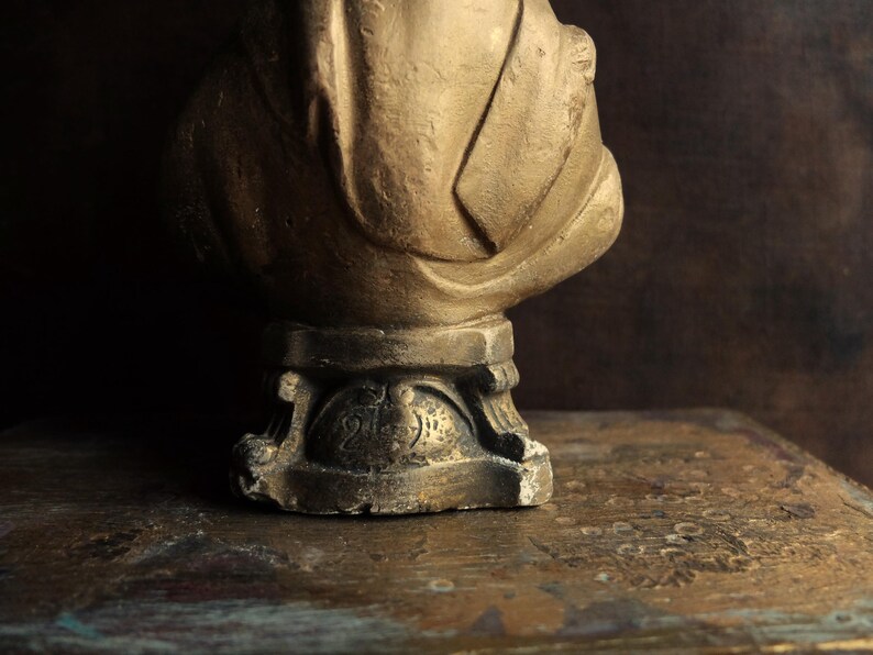 May include: A gold-colored statue of a person with a draped cloth, standing on a pedestal with a decorative base.