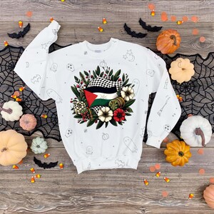 Op de afbeelding: Witte trui met een zwart-wit geblokte vlag met groene, rode en zwarte accenten, omringd door bloemen en bladeren. De trui heeft een Halloween-thema met kleine zwart-witte illustraties van geesten, vleermuizen en pompoenen.