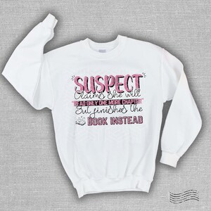 May include: White sweatshirt with the text "SUSPECT Claims she will READ ONLY ONE MORE CHAPTER But finishes the BOOK INSTEAD" in pink and black lettering. The sweatshirt has long sleeves and a crew neck.