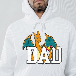 May include: White hooded sweatshirt featuring a cartoon dragon with orange, teal, and yellow accents. The word "DAD" is printed in bold, white letters with a black outline. The design is centered on the front of the sweatshirt.