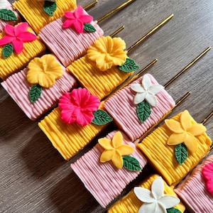 May include: An overhead shot of several rectangular treats on gold sticks, decorated with colorful flowers and green leaves. The treats alternate between yellow and pink, with various flower colors including pink, yellow, and white.
