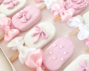 May include: A box of decorative, pastel-colored treats on wooden sticks. The treats are oval-shaped, with pink and white icing, adorned with bows and decorative details. Some have pearl accents.