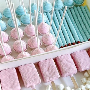 May include: A box of assorted treats, including pink and blue cake pops, pink ice cream bars on sticks, and blue-coated pretzel rods. The cake pops have a spiral design, and the ice cream bars have a textured surface. White marshmallows are scattered around the box.