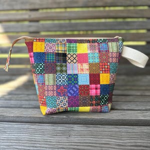 May include: A colorful patchwork fabric bag with a variety of patterned squares in shades of yellow, blue, pink, and green. The bag has a beige canvas handle on each side and a beige interior. The bag is sitting on a wooden surface.