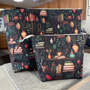 May include: Two zippered pouches with a black background and a repeating pattern of books, plants, and vases. The larger pouch is on the bottom and the smaller pouch is on top. Both pouches have a white zipper.