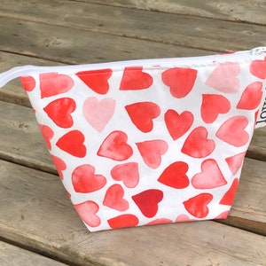 May include: White zippered pouch with a red and pink watercolor heart pattern. The pouch has a white label with the word "loved." printed in black.