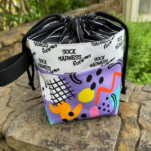 May include: A purple drawstring bag with a black handle and a colorful geometric pattern. The bag has a white lining with the text "Sock Madness Forever" printed on it.