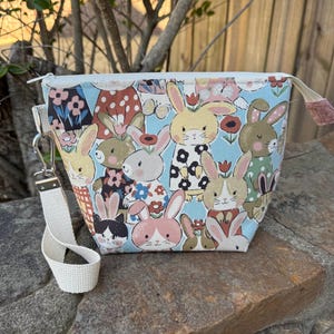 May include: A small, square fabric pouch with a zipper closure and a white strap. The pouch is decorated with a repeating pattern of cartoon bunnies in various colors and outfits against a light blue background. The strap is attached with a silver-colored clasp.