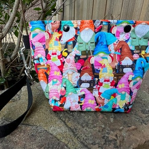 May include: A black and white fabric tote bag with a colorful print of gnomes wearing hats and holding sewing tools. The bag has a black strap with a silver clip.
