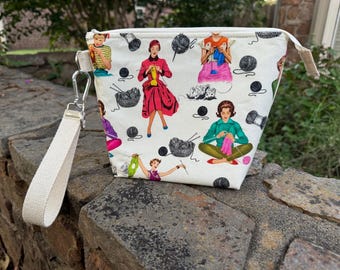May include: White zippered pouch with a colorful print of women knitting. The print features women in various poses, yarn balls, knitting needles, and a basket of yarn. The pouch has a white strap.