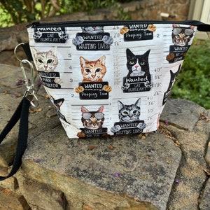May include: A white zippered pouch with a black handle. The pouch is decorated with a pattern of cats wearing sunglasses and holding signs that say "Wanted" with various crimes listed, such as "Cat Burglary" and "Wasting Lives".