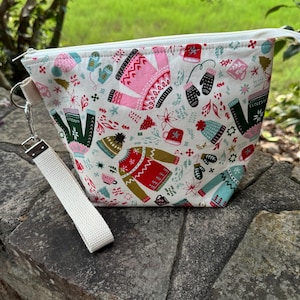 Puede incluir: Una bolsa blanca con cremallera y correa para la muñeca, adornada con un estampado festivo de suéteres, mitones y accesorios de invierno en rosa, verde, rojo y azul. La bolsa está sobre una superficie de piedra.