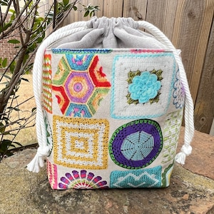 May include: A colorful drawstring bag with a patchwork design. The bag features various crochet squares in vibrant colors like red, yellow, blue, and green. It has a gray top and white rope drawstring.