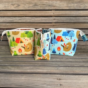 May include: Three fabric pouches with a camping theme. The pouches feature a repeating pattern of bears, tents, trees, and campfires. The pouches are in shades of tan, blue, and green, with white zippers and plaid accents.