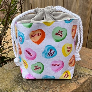 May include: White drawstring bag with a white cotton lining. The bag is printed with colorful candy hearts that say "Love You", "Kiss Me", "Be Mine", "Sweet", and "XOXO".