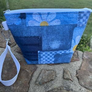 Puede incluir: Una bolsa de mezclilla azul con cremallera blanca y correa de muñeca. La bolsa presenta un diseño de patchwork con patrones de margaritas y cuadros. La correa de muñeca está sujeta con un mosquetón plateado.